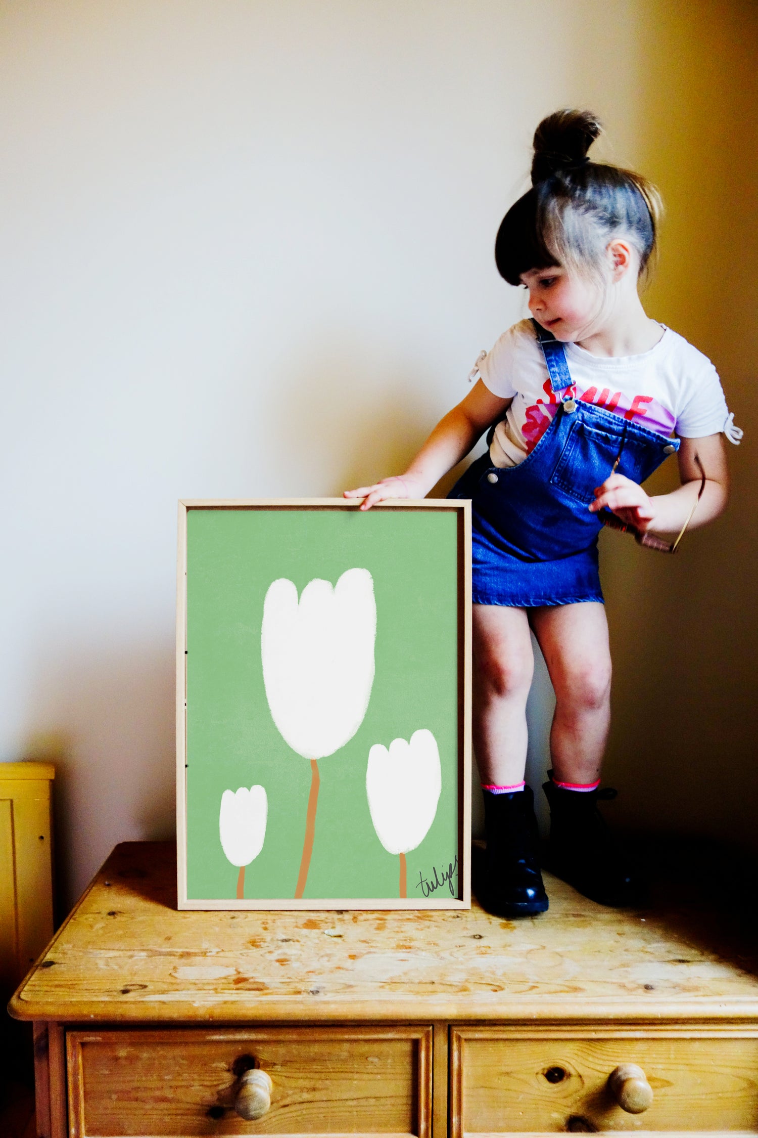 small girl stood on drawers looking at framed picture of hand drawn tulips in white on green background